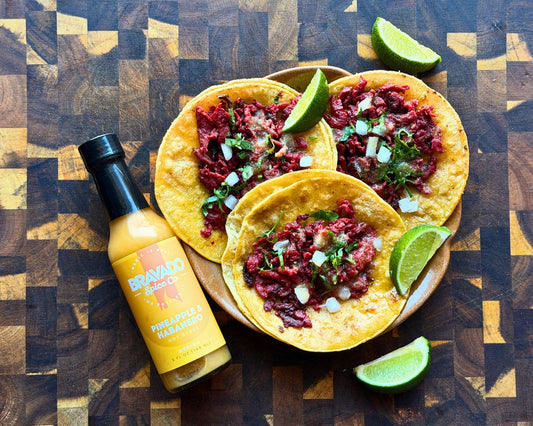 Three street tacos with meat, cilantro, onions, and limes next to a bottle of Pineapple Habanero sauce.
