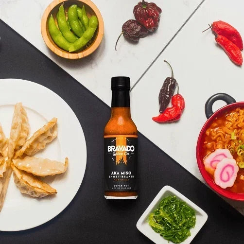 A bottle of Bravado Spice AKA Miso Ghost-Reaper hot sauce beside chili peppers, ramen, and dumplings.