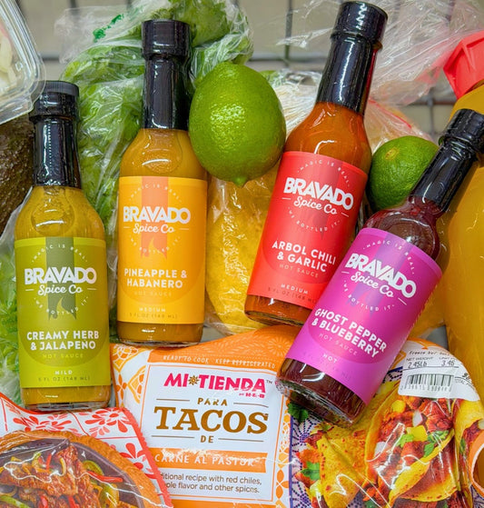 Four Bravado Spice hot sauce bottles displayed with taco ingredients and fresh limes.