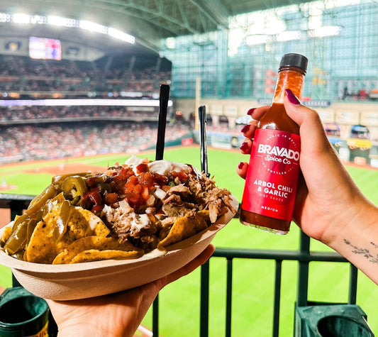 A hand holds tacos in one hand and Bravado Spice Co.’s Arbol Chili & Garlic hot sauce in another on a Game Day.