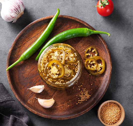 Sliced green jalapeños and mustard seeds submerged in a clear fermentation brine within a glass jar.