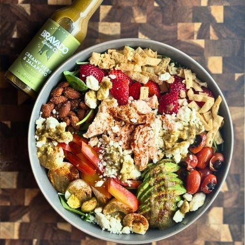 Colorful salad with strawberries, nuts, and a bottle of Bravado hot sauce on a wooden surface.