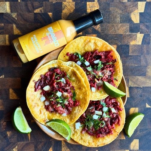 Three tacos on wooden cutting board with Pineapple and Habanero Hot Sauce with limes to garnish