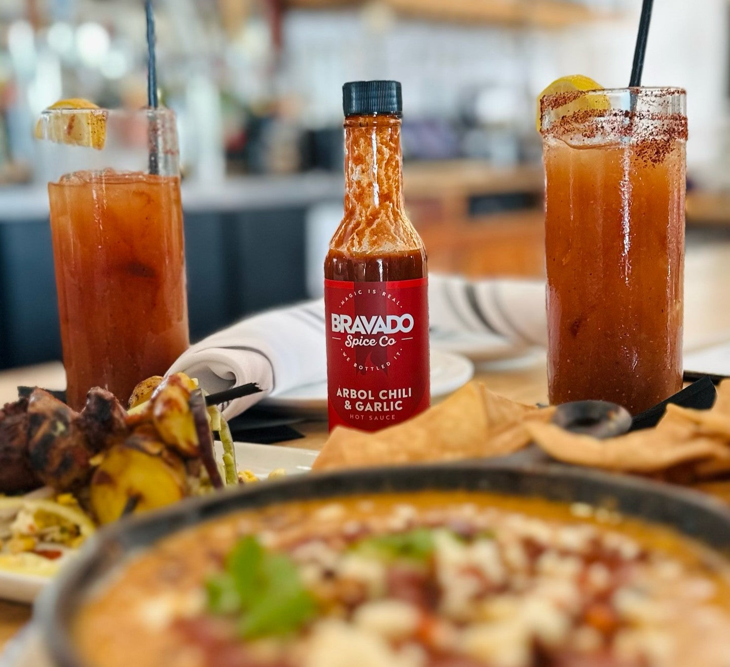 Bottle of Bravado Spice Arbol Chili & Garlic hot sauce on a restaurant table surrounded by cocktails, chips, and plated dishes.