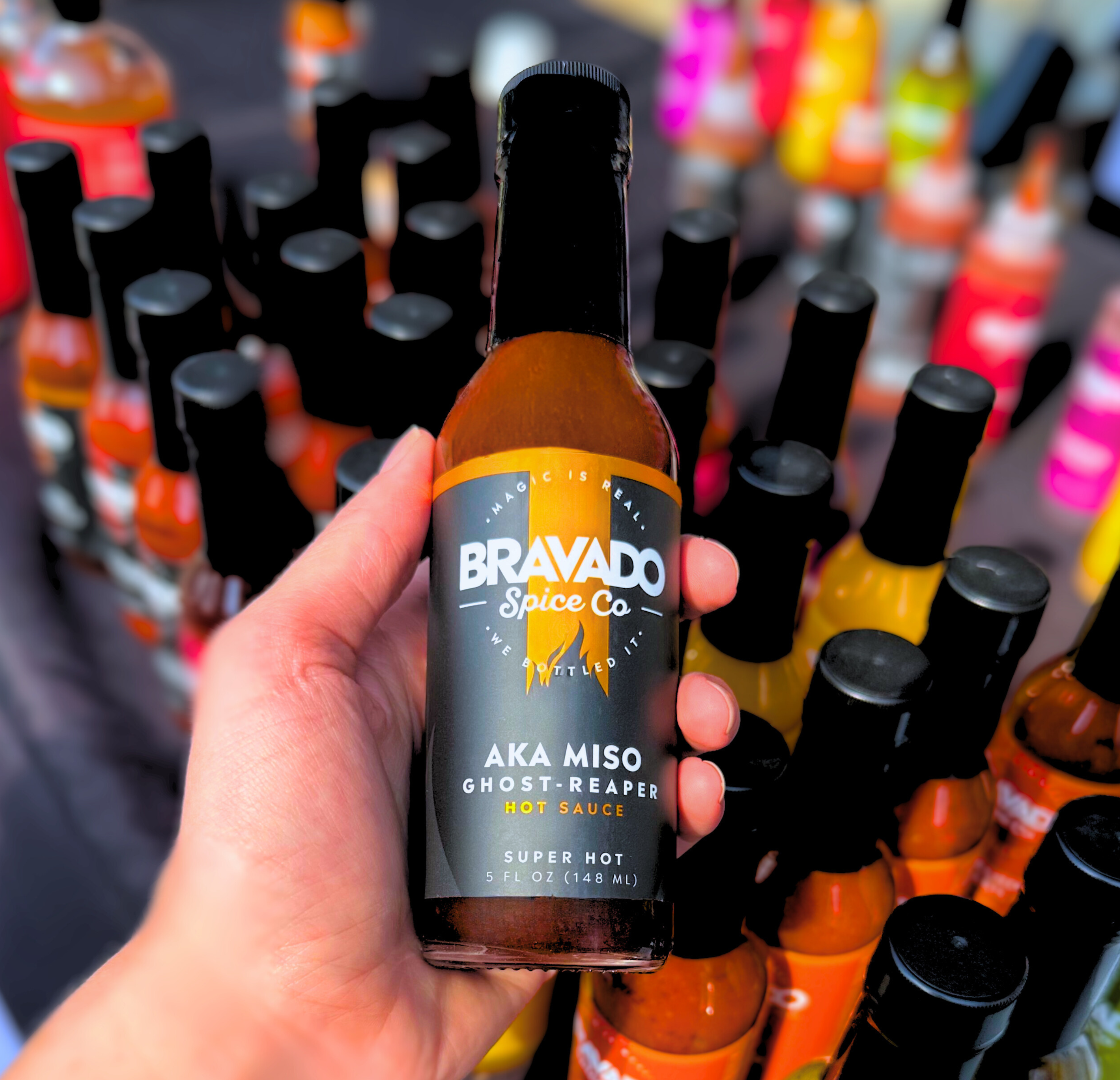 Hand holding a bottle of Bravado Spice Co Aka Miso Ghost Reaper hot sauce in the foreground, with rows of colorful Bravado hot sauce bottles blurred in the background at a display or event setup.